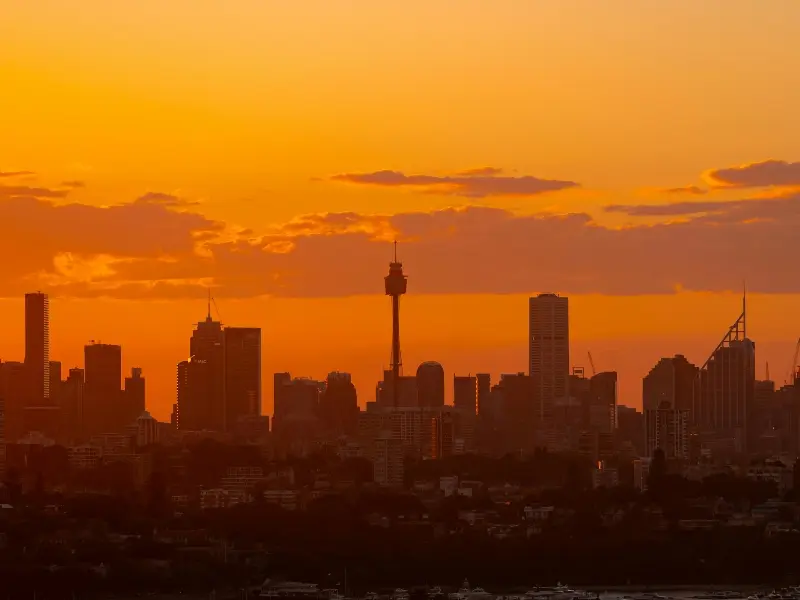 Sydney city skyline at sunset with an orange sky and silhouetted high-rise buildings, including the Sydney Tower.