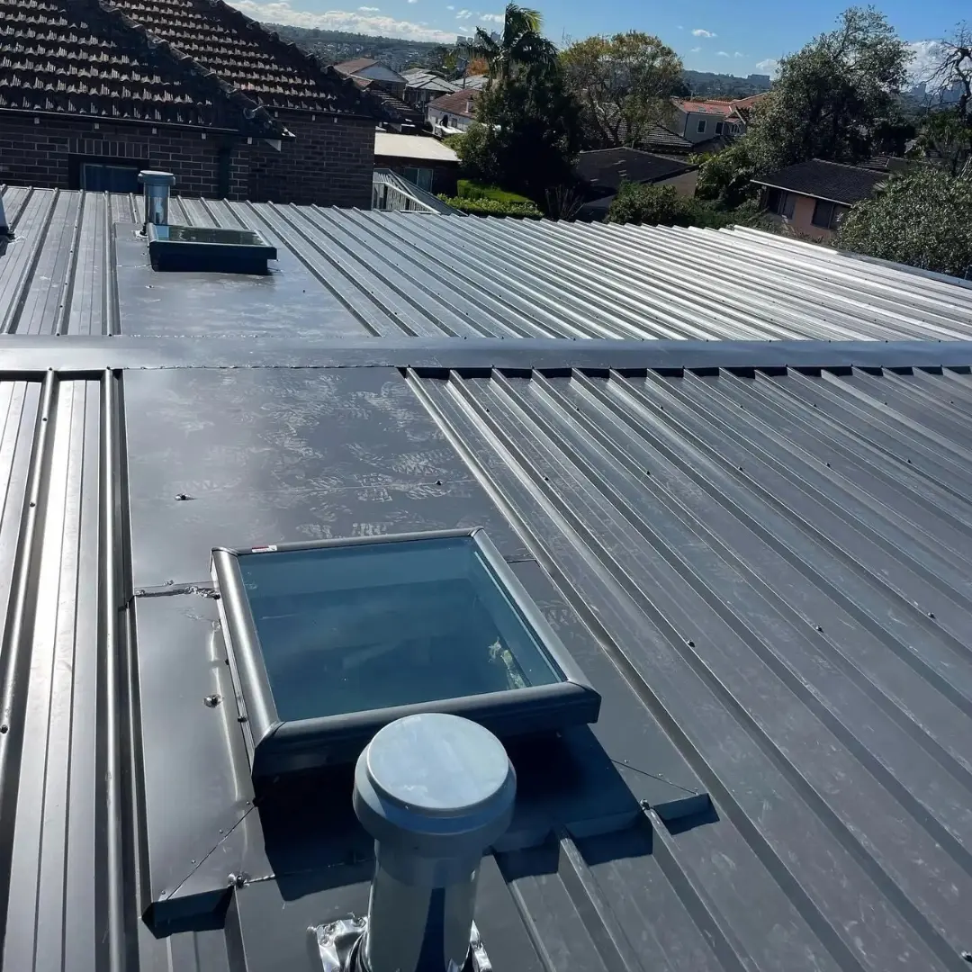 Rectangular skylight installed on a corrugated metal roof with a nearby roof ventilation pipe on a residential property.